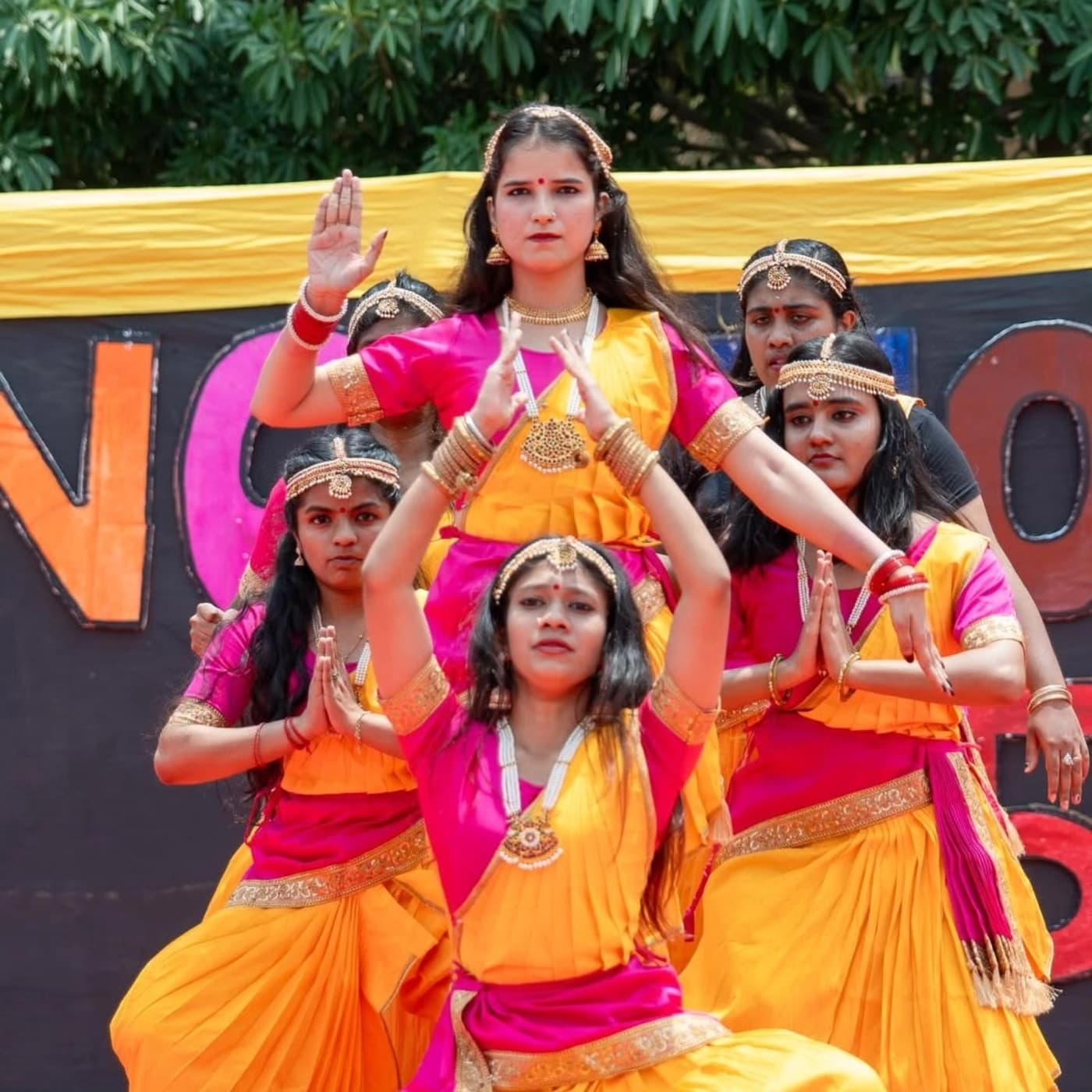 A classical dance troupe performing in pink and gold costume on an outdoor stage