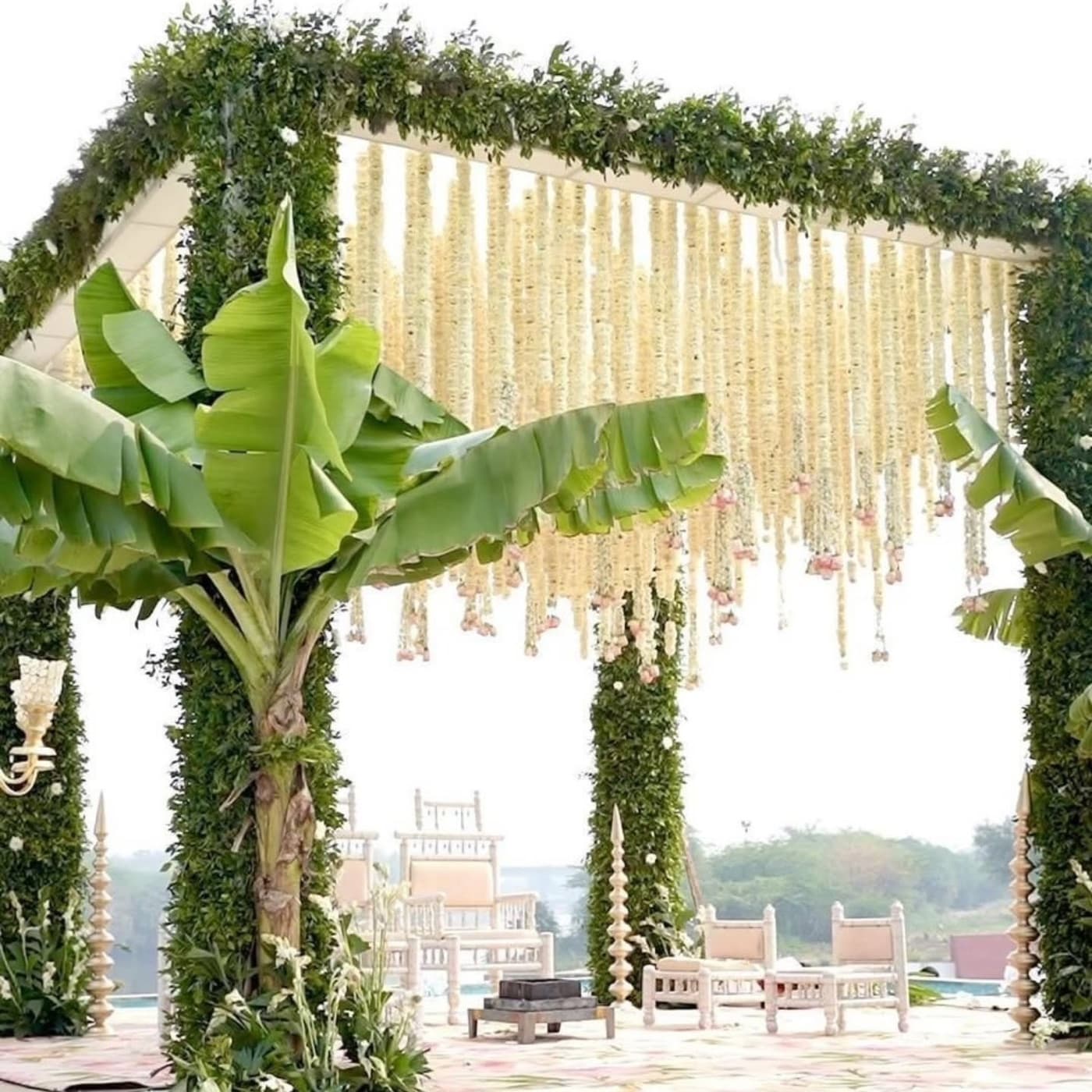 An open-air mandap wrapped in foliage with cascading jasmine strands beside a pool