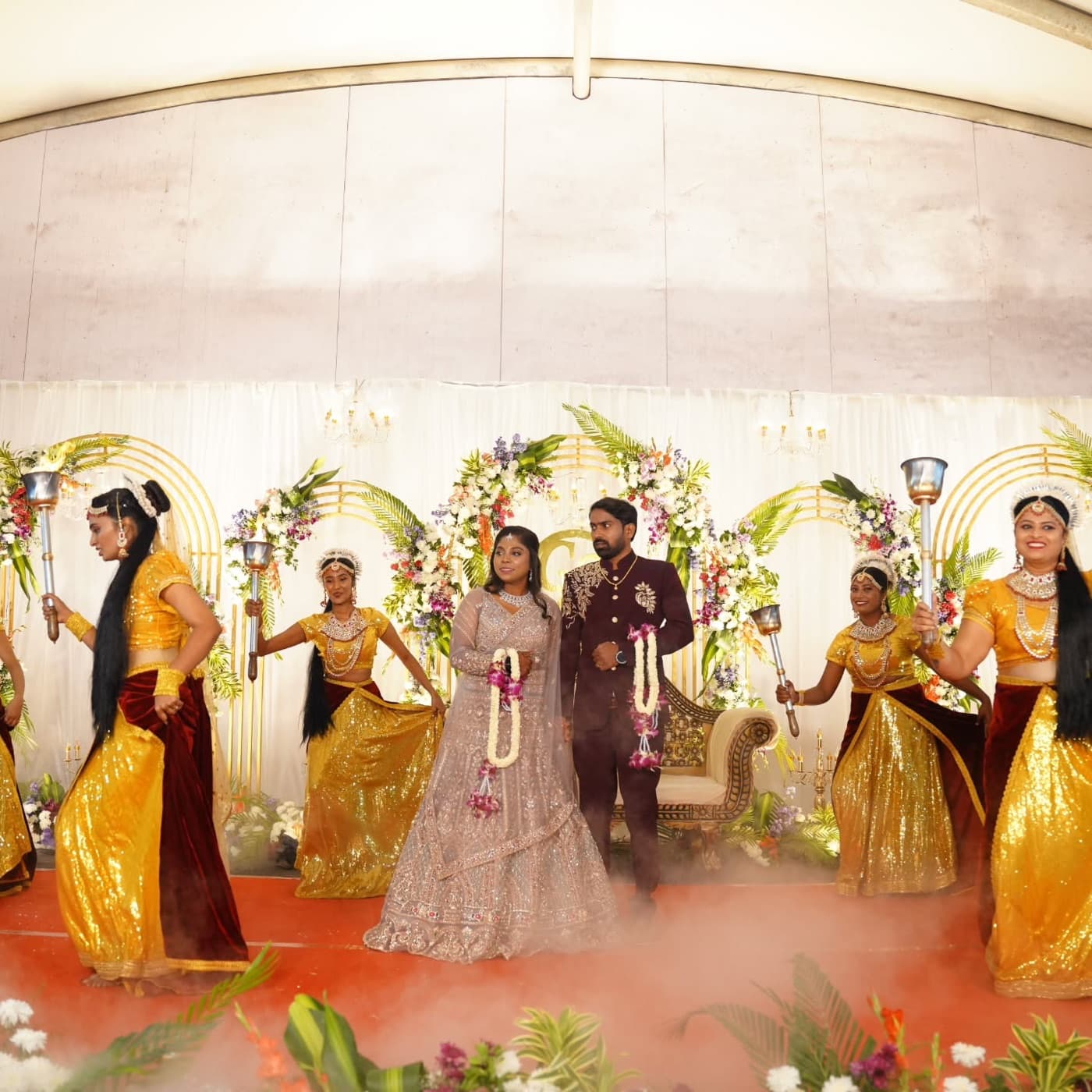 A couple entering a sangeet stage flanked by dancers holding lit torches