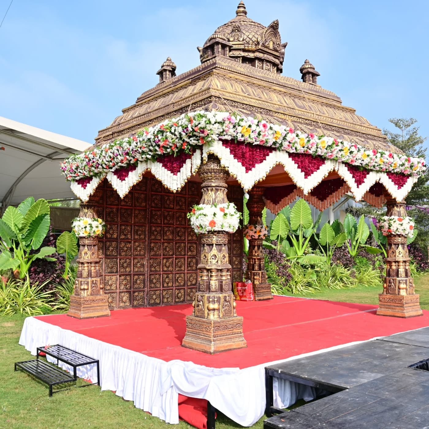 A carved wooden temple-style mandap edged with white and magenta garlands on a lawn